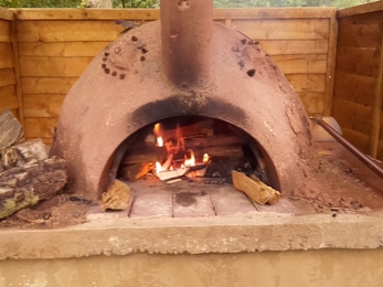 Clay pizza oven with fire inside and fence panels surrounding it.