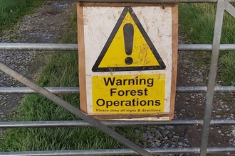 Yellow warning sign on gate across track bordered by hedgerow