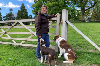 Woman stood by gate in field with two brown and white dogs sat by her feet