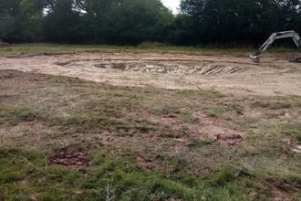 Shallow hole with digger arm visible on right hand side of image, grassy around and trees in the background