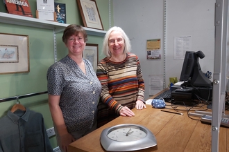 Two ladies stood by cash desk smiling at camera