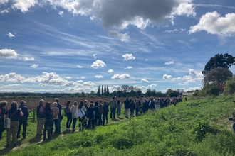 Long line of people along a path through open grassland with blue sky and fluffy white clouds above
