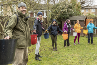 a group of volunteers stand in a garden in a line, they are all wearing waterproofs and wet weather gear. they are passing buckets along their line and smiling at the camera. 