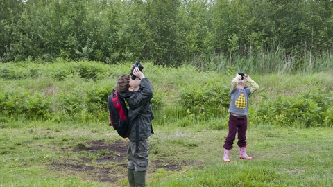 Two children in a grassy outdoor area looking up with binoculars