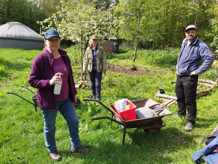 Three people stood, looking at the camera, with a wheelbarrow and tools in a garden
