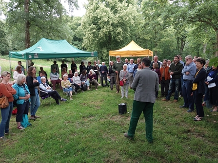 Audience of people stood around a man, outdoors, a green and orange gazebo in background