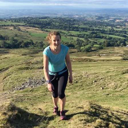 Woman in pale blue T Shirt walking up a hill with view of landscape beyond