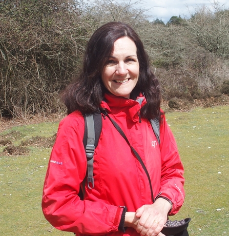 Woman in red coat with long dark hair smiling at camera in countryside setting