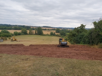 Digger moving earth attop of sloping field with thick hedgerow on right hand side