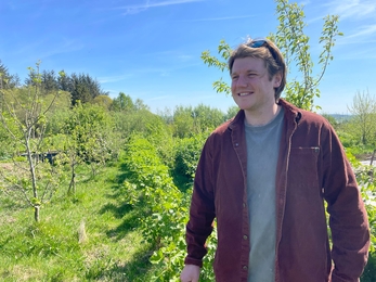 Man in dark red shirt stood outdoors in front of young trees in rural scene