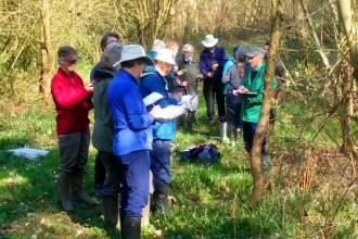 Group of people in grassy, wooded area, taking notes