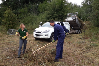 Man and woman raking straw in a field with a white vehicle in the background