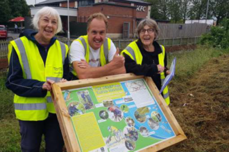Three people in hi-vis jackets standing behind an interpretation board smiling at ca,era