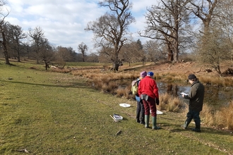 Three people stood by pond in grassland dotted with trees; winter