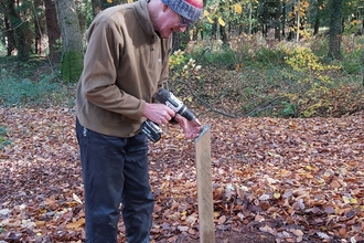 Volunteer Paul is drilling a Tree description plate to the top of a wooden post