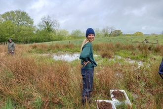 Woman in blue beanie turning to smile at camera, stood in grassy field in front of pond