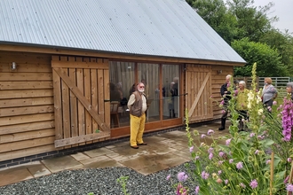 Ban stood in front of barn with wooden sides and sloping grey roof with flowers in foreground on right hand side.