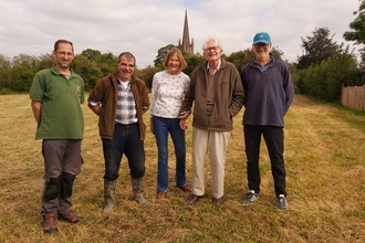 Four men and one woman stood in a line facing the camera, smiling, in a grassy field with church spire in the background