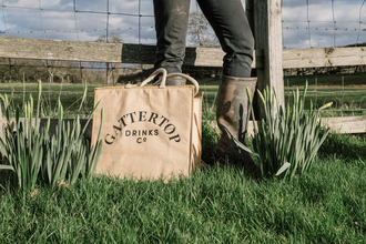 Jute bag in front of a person, legs only visible, in front of fence, surrounded by daffodils.