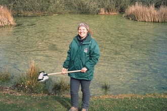 Woman with net on a stick stood in front of a pond wearing a green coat