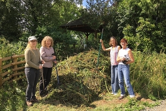 Four women holding tools stood by a large pile of cut grass