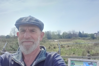 Man wearing a cap looking at camera  with notice board and wild area of grass, water and trees behind