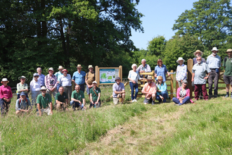 About two dozen people stood and kneeling for a group photo in a field in the sunshine with a notice board in the centre