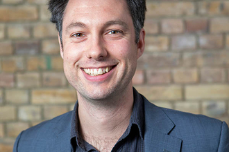 Headshot prtrait of man with dark hair wearing a blue suit jacket and open shirt smiling at the camera