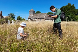 A man stood up and a woman knelt down facing each other in long grass in front of a small church in late summer