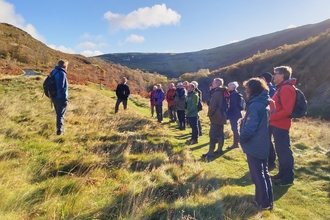 Group of people wearing outdoor coats and rucksacks on grass with mountains beyond