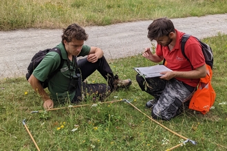 Two men sat on grass by a square wooden frame - one with clipboard