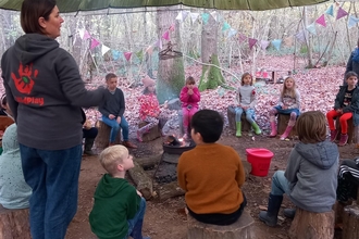 Group of children sitting around a fire on tree stumps under a canopy 