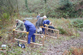 A group of volunteers dressed in dark blue waterproofs are workign with wood to make a bridge with handrails. They are in the forest adn there are autumn leaves on the ground