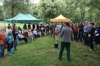 Audience of people stood around a man, outdoors, a green and orange gazebo in background