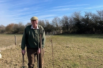 Man in wellies and flat cap stood in field with young tree saplings and tree guard