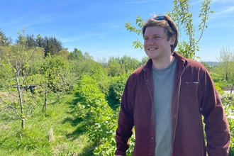 Man in dark red shirt stood outdoors in front of young trees in rural scene