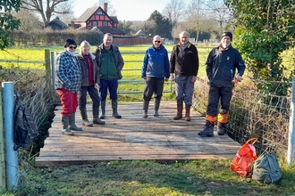 Six people in outdoor clothes stood on a flat wooden bridge with field visible behind