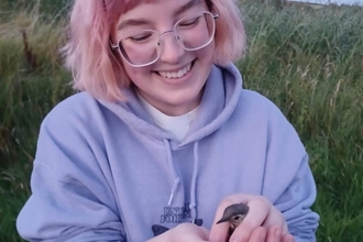 Young woman with pink hair and wearing a mauve hoodie holding a small bird in cupped hands with grassland in background