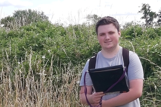 Young man holding a black folder stood outdoor with hedgerow behind