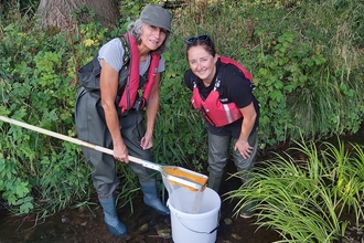Two women stood in stream with white bucket and net, smiling at camera