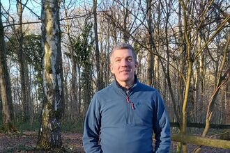 Man wearing a blue jacket stood facing camera in a woodland