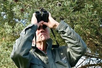 Man in dark green coat looking upwards through binoculars