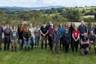 Around 25 people stood on a lawn for a group photo with rural landscape in background