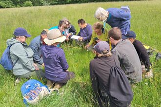 Small group of about 10 people sat in a rough circle in a meadow of long grass