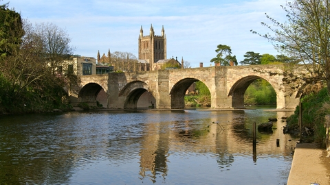 View of wide river flowing under 4 arches of a stone bridge with cathedral tower behind