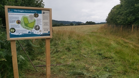 Information board with map at entrance to grassland bordered by trees and hedges