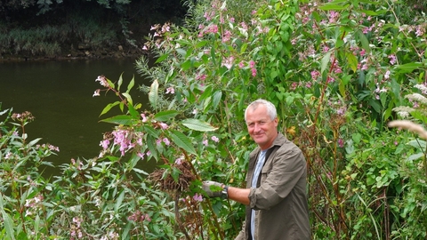 Man stood beside huge plant with pink flowers, a river visible in background
