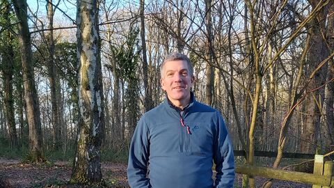 Man wearing a blue jacket stood facing camera in a woodland