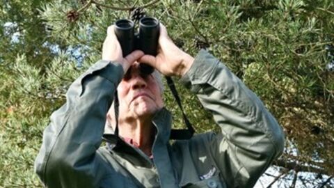 Man in dark green coat looking upwards through binoculars