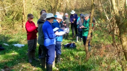 Group of people in grassy, wooded area, taking notes